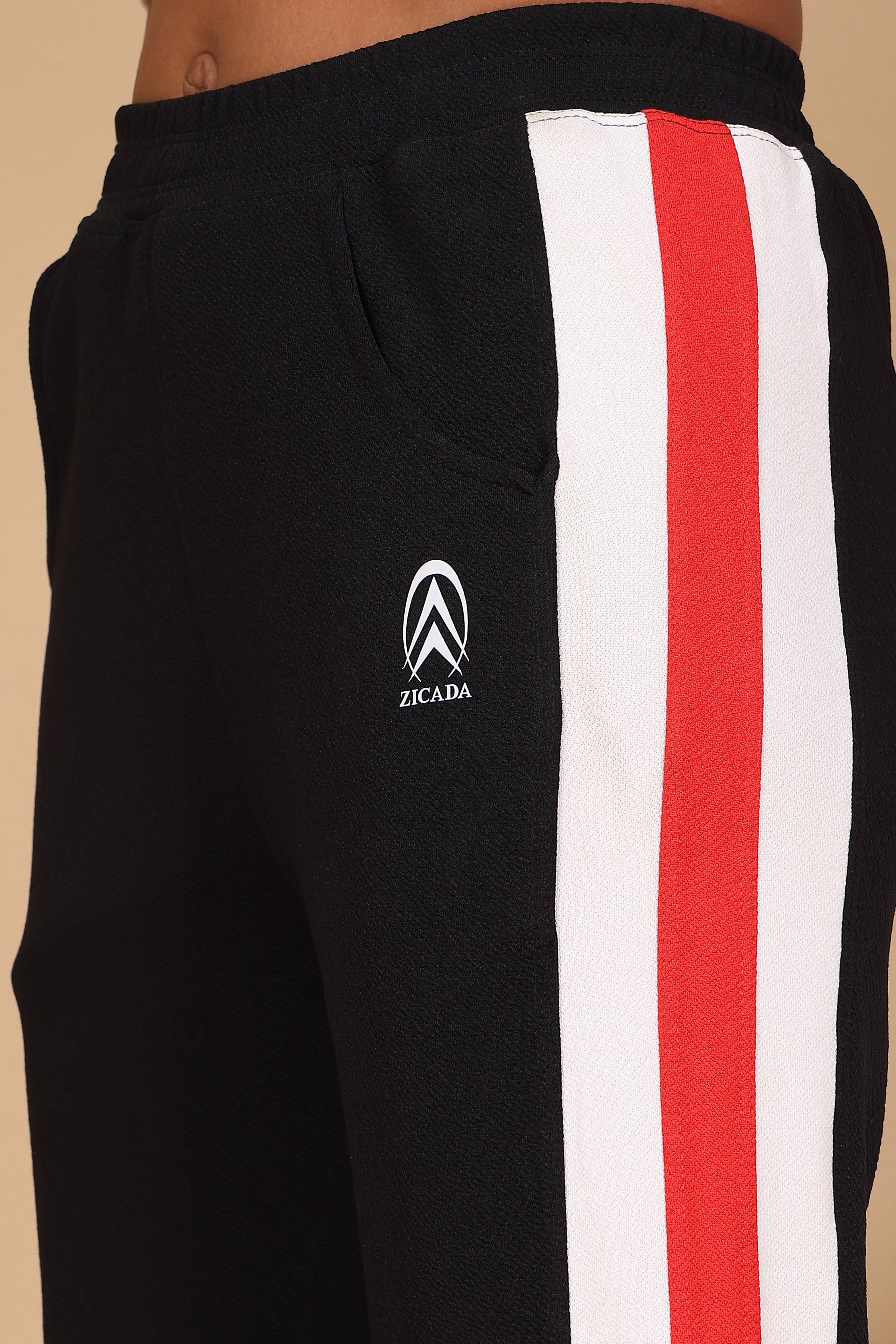 Black sweatpants with red and white stripes and a ZICADA logo on a beige background