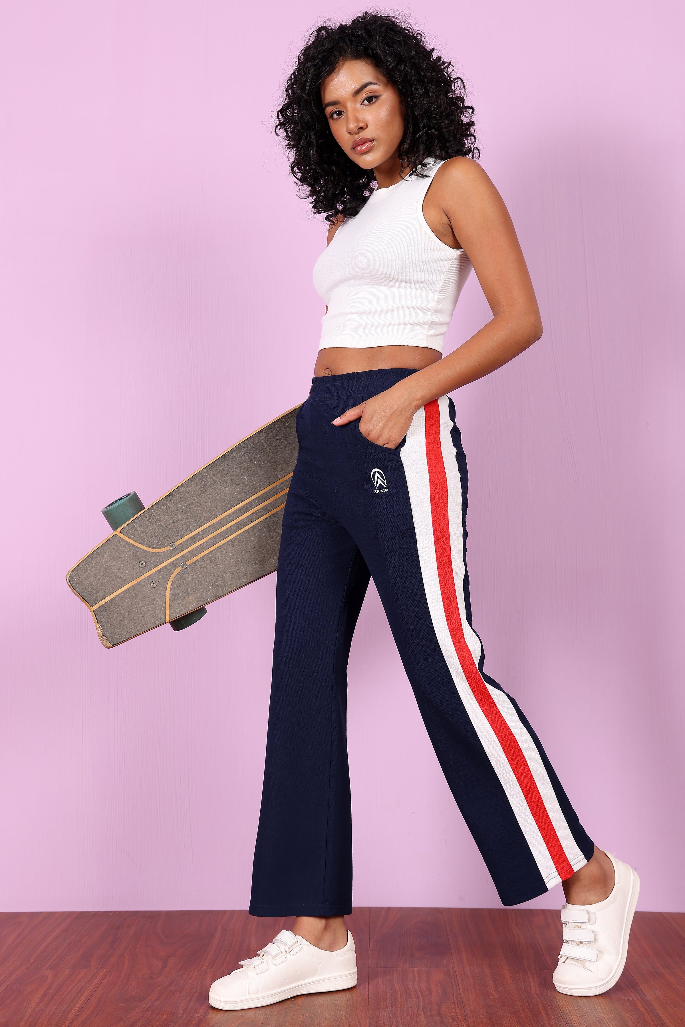 Woman holding a skateboard and Wears a ZCIADA wearing navy pants with red and white stripes against a pink background