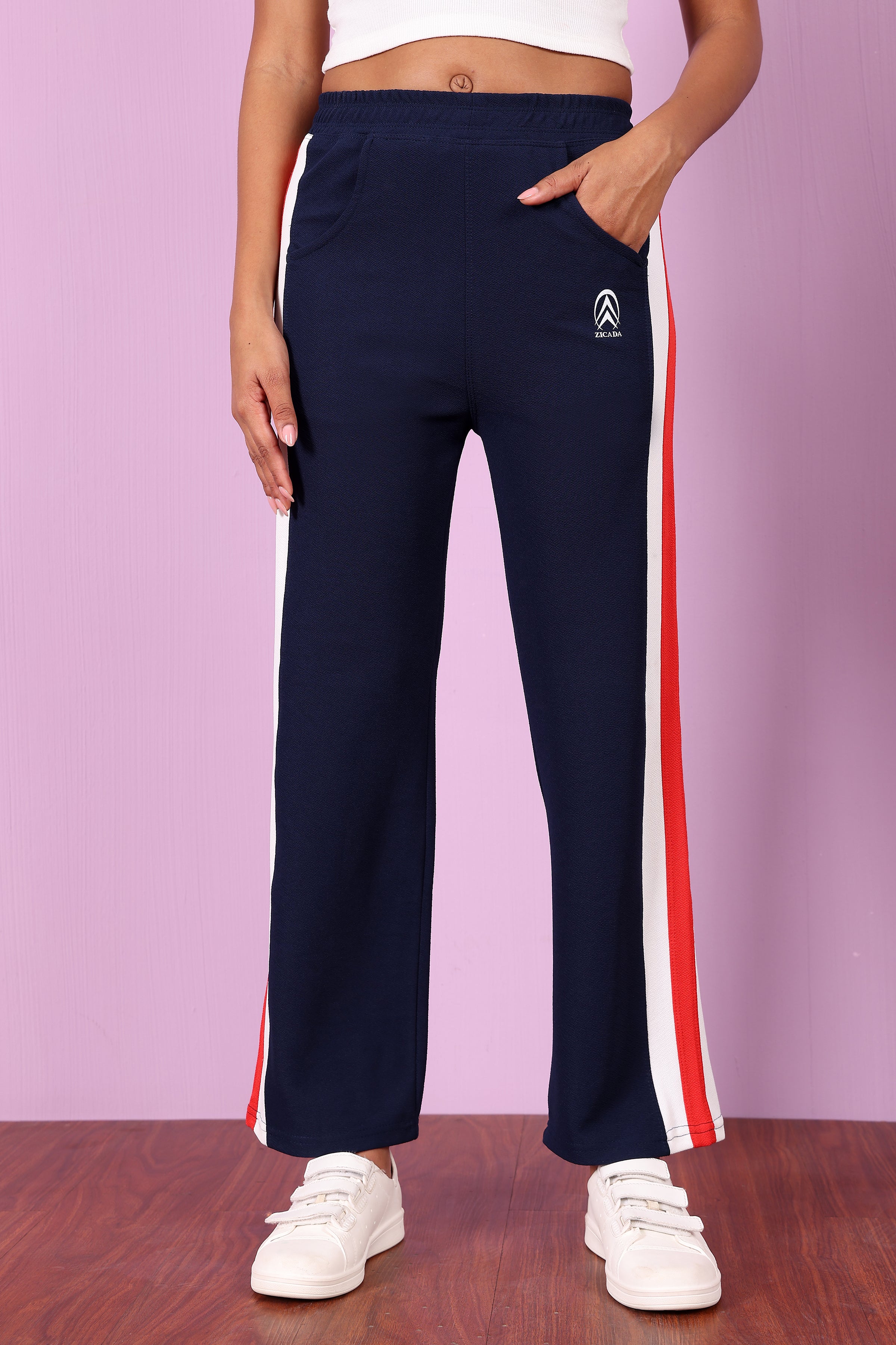 Zicada Navy track pants with red and white stripes on a wooden floor and pink wall background
